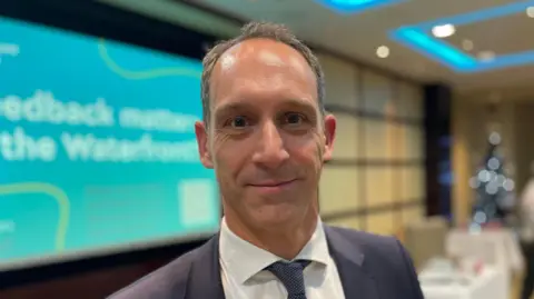 BBC Lee Henry is a middle-aged white man with short grey hair. He is smiling into the camera and wearing a navy suit with a white shirt and blue tie. The background is an out-of-focus meeting room with a blue projection on a screen.
