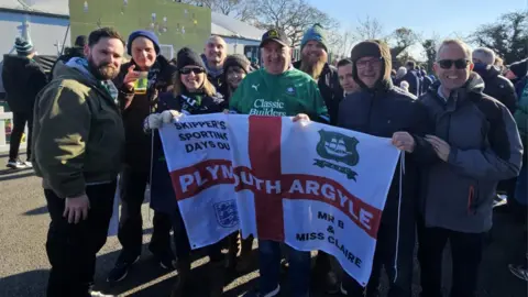 A group of Plymouth Argyle supporters from the North Devon Greens group stand outside a football stadium holding an England flag with the team's name, the England national team badge, Argyle badge and messages on it. A big screen showing another football match is in the background.