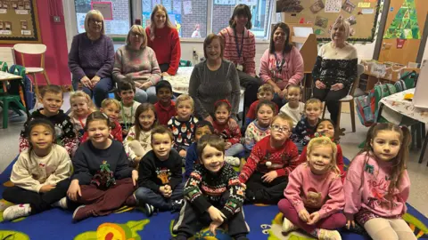 About 20 primary school age children sit on the floor. behind them six adults sit on chairs