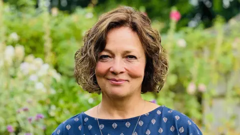 Branwen Jeffreys/BBC Professor Julia Waters standing in front of garden foliage. She is smiling and has a light brown wavy-haired bob and is wearing a teal blue top patterned with a light leaf motif