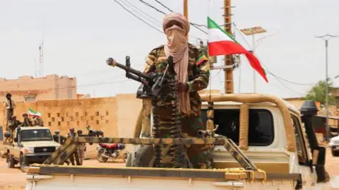 AFP An armed rebel fighter in military fatigues stands on the back of a pick-up vehicle in a parade. A flag is attached to the vehicle.