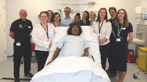 Bradford Teaching Hospitals Ten people stand around a hospital bed with a pregnant manikin lying in it