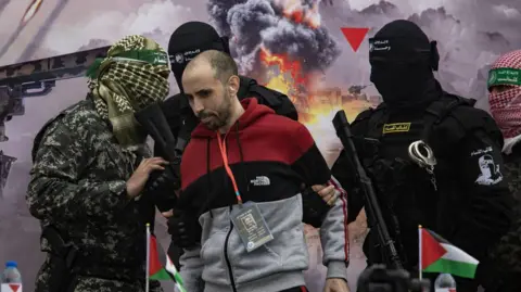 EPA-EFE/REX/Shutterstock Tal Shoham on stage before gunmen handed him to the Red Cross in February. Men with their faces covered and holding guns stand either side of him.