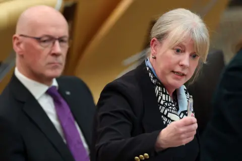 Getty Images Shona Robison, a woman with blonde hair tied back, is pointing with a pen as she answers a question in the Scottish Parliament chamber. She is wearing a black jacket and black and white scarf. John Swinney, a bald man with glasses, is sitting behind her. He is wearing a black jacket, white shirt and purple tie