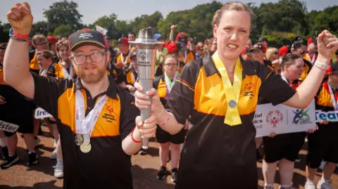 Jenna Thevanau (right) holding a Special Olympics flame with another male athlete. Both are holding their other hand in the air.