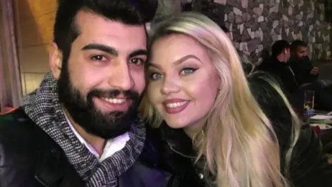 A couple smile at the camera. She has blonde hair and blue eyes. He has dark hair and a beard, and brown eyes. They are taking a selfie inside a restaurant or bar.