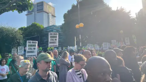 Crowds walking with placards saying 'this much evidence and still no charges'. In the background is Grenfell Tower