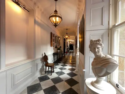 Long, elegant hallway with white panelled walls and a black-and-white checkered floor, lit by hanging lantern-style lights, featuring a marble bust by a window, framed artwork, and a small table with chair along the wall.