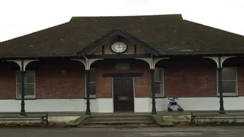 Camberley and District Shed the outside of a long single-storey sports pavilion, with slim sash windows and steps leading up to the door under a covered porch with Victorian-style pillars and a clock above the door