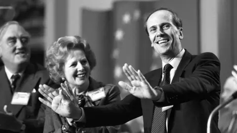 Margaret Thatcher with Norman Tebbit as he appeals to delegates to stop the applause after his speech at the opening of the annual Tory conference at the Winter Gardens, Blackpool. He is seen in a dark suit with a dotted tie.