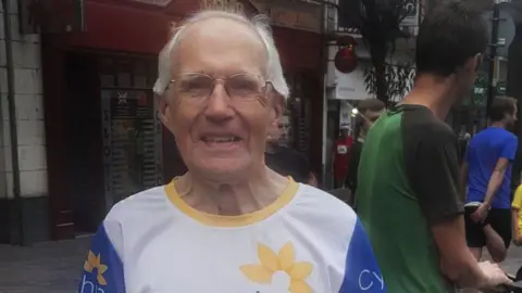 Trevor Hardless with short white hair wearing a white running outfit. He is standing by railings in Northampton town centre. There are people walking past and one man standing by a bike.