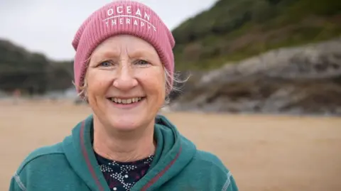 Carol Maddock stands on a beach smiling at the camera. She is wearing a green hoodie and a pink beanie hat that says "ocean therapy" 