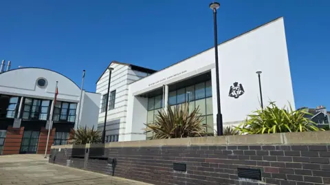 The outside of the Courts of Justice, which is a large, white, glass-fronted building with the government crest on the right. The photograph is taken from behind a long dark stone wall with plants lining it. There are unbroken blue skies.