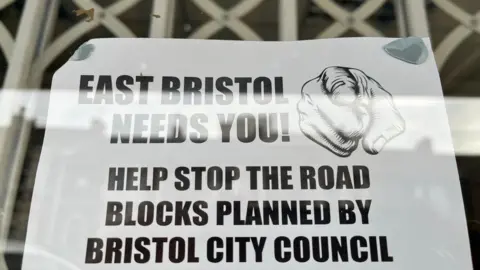 A black and white sign on a piece of paper in the window of a cafe on Avonvale Road asks for people to protest against the East Bristol LTN trial.