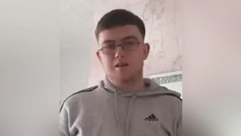 Daniel Ambler, a 23-year-old man with short dark hair, wearing spectacles and a grey hoodie. He is standing in what looks like a living room. There is pink and grey wallpaper on the wall behind him.
