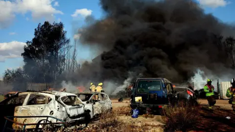 South Yorkshire Fire & Rescue Firefighters tackling a large fire behind a chain-link fence. In the foreground, burnt out cars can be seen.