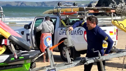 A lifeguard in a wetsuit with a rash vest over it is turning a metal winch which is lowering a jetski off its trailer. His face is contorted with effort. He is on a beach with the sea behind him on a sunny day. There is a RNLI-branded pick-up truck behind him which is loaded with paddleboards. A man with a towel around his waist and wearing a hoodie is stood in the open passenger door way.