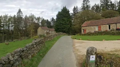 Google A lane in Urra following a dry stone wall with stone houses and outbuildings on either side.