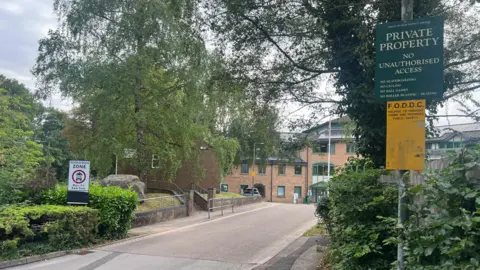 Carmelo Garcia A wide shot of the Forest of Dean District Council building, a red-brown brick office block in the background. The driveway leads toward the entrance, and a sign on the right reads: “Private property, no unauthorised access.”