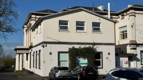 A close-up of a large white building with a wing to the right. There is a sign on the front that reads "Lillington House", and it has a green bush growing around it. There are cars parked outside, and on the left-hand side of the building, there is an entry with a porch and pillars.