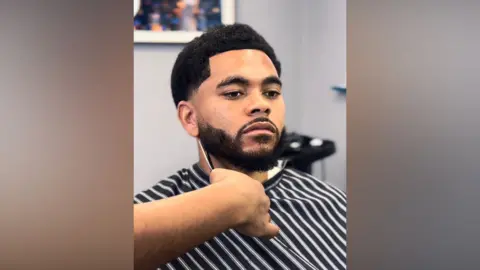 Musaab Somobwane A man wearing a barber cape sits in a chair, showing a close‑up of a haircut.
