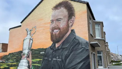 BBC A wall mural showing Shane Lowry wearing a dark Bank of Ireland polo shirt and smiling while holding a silver trophy