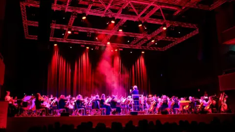 City of Wolverhampton Council The full orchestra is bathed in deep red lighting with a conductor standing on a podium at the centre. Smoke rises through the red light while musicians play string, wind and percussion instruments across the wide stage. Audience members' heads are silhouetted in the foreground.