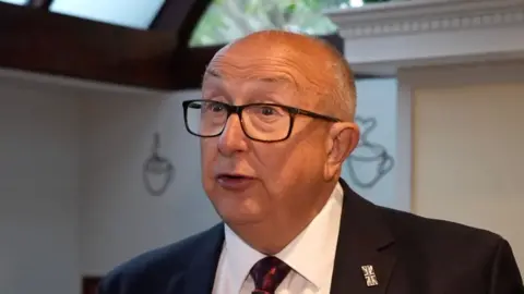 Andrew Sinclair/BBC A man wearing black glasses and is balding looks towards the camera while wearing a suit and tie inside a white room with a skylight