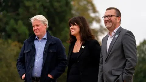 PA Media Earl Spencer with short white hair, a blue jacket and blue shirt, with his hands in his pockets, Caroline Redman Lusher with long dark hair and a black top and jacket, and Nigel Varndell with short dark hair and beard, wearing a grey suit with his hands in his pockets. There are trees behind them.