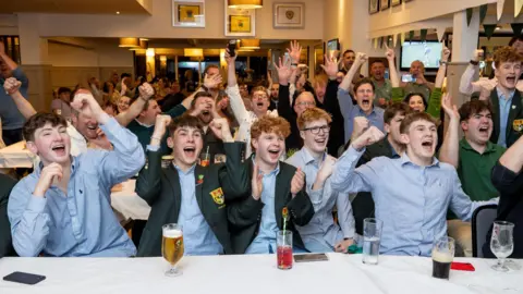 A group of young people sit at the front of a large crowd in a golf club supporting Rory McIlroy. They are all wearing blue shirts and the two young men in the middle are also wearing dark green golf club blazers. Drinks sit on the table. The people are all cheering and punching the air.