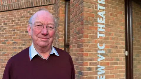 BBC John Sambrook, a man wearing glasses and a maroon coloured jumper. He is standing next to a brick-building with white lettering on the side which reads New Vic Theatre.