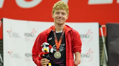 Para-badminton athlete Rob Donald stands proud with a silver medal around his neck. He is smiling and holding what appears to be a dog mascot, that is saluting, in one hand. 