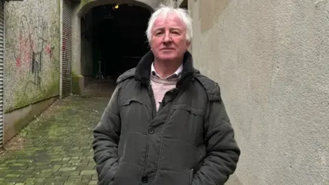 Noel is standing at an entry with his hands in his pockets.