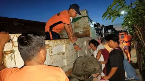 A man pulls a bag up onto a truck, while a woman leans her head against the side of the truck