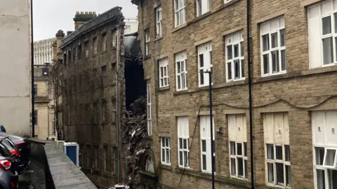 A long line of white-framed windows in a five-storey building with a sudden gap where the wall has fallen away