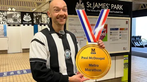 Paul McDougal stands in front of St James' Park Metro station wearing Newcastle football black-and-white striped kit. He is holding up a large paper medallion which says he has completed a Metro marathon and credits his name. Paul has a shaved head and stubble. 