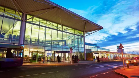 The exterior of the main terminal building at Bristol Airport. It is a large, modern structure with full-length glass windows, and the picture has been taken at dusk, with a dark blue, slightly cloudy sky. Passengers can be seen entering the terminal. 