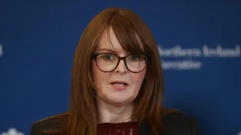 PA Media Caoimhe Archibald  has auburn hair and glasses standing in front of a blue background
