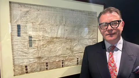 Gavin Kermack / BBC A man with short brown hair and black-rimmed glasses is wearing a red tie, pale shirt and dark blue jacket. He is standing in front of a very old document in a glass frame, the details of which cannot be read.