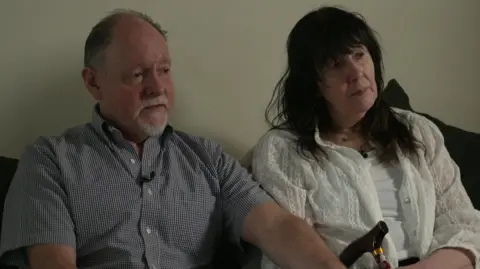 A man in a check shirt is sitting next to a woman who is holding a walking stick. His hand is on her hand.