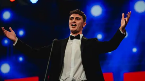 Celf Calon Caleb Nicholas stands on stage wearing a black suit and white shirt. His hands are outstretched as he sings. He has short dark hair. 
