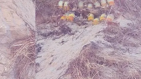 Several SIPs (self-igniting phosphorus) explosives in yellow/orange shaded bottles are laid next to each other in beach sand. All of the items have a yellow/orange band around the bottles. There is grass among the sand.