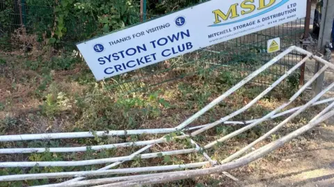Syston Town Cricket Club The entrance to Syston Town Cricket Club shows a flattened gate and a hanging sign. 