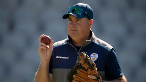 Derbyshire head of cricket Mickey Arthur wearing a baseball glove and holding a cricket ball in the other hand