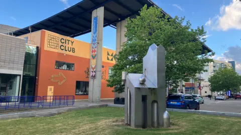 Amy Holmes/BBC Modern city plaza with a bright orange “City Club” building, a tall steel canopy on concrete pillars, a geometric metal sculpture, and trees lining a quiet street under a clear blue sky.
