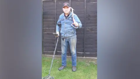 Noonans Tom Clarke in double denim and holding his metal detector in his right hand, his spade in his left