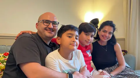 Akmal is wearing a dark grey top and wearing black square glasses, Gafur is wearing a white t-shirt, Bobur is wearing a red t-shirt and Gavhar is wearing a black vested top and a black and white checkered skirt and sitting on a blue sofa. 