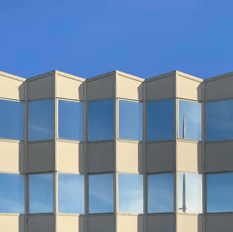 Tom Hicks Blue sky is reflected in the windows of a white building with shadows making it look corrugated. 