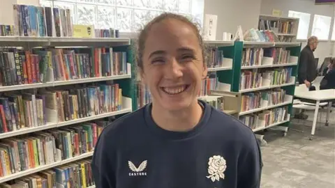 James Burridge/BBC Abby Dow smiling at the camera in a navy England rugby shirt. She is standing in a library.