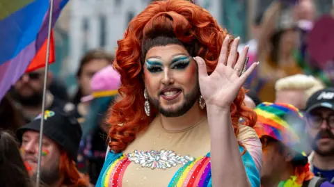 Matthew Horwood/Pride Cymru A person wearing make up, with a dark beard and long red hair wears a pink top with rainbow colours as they wave to the camera